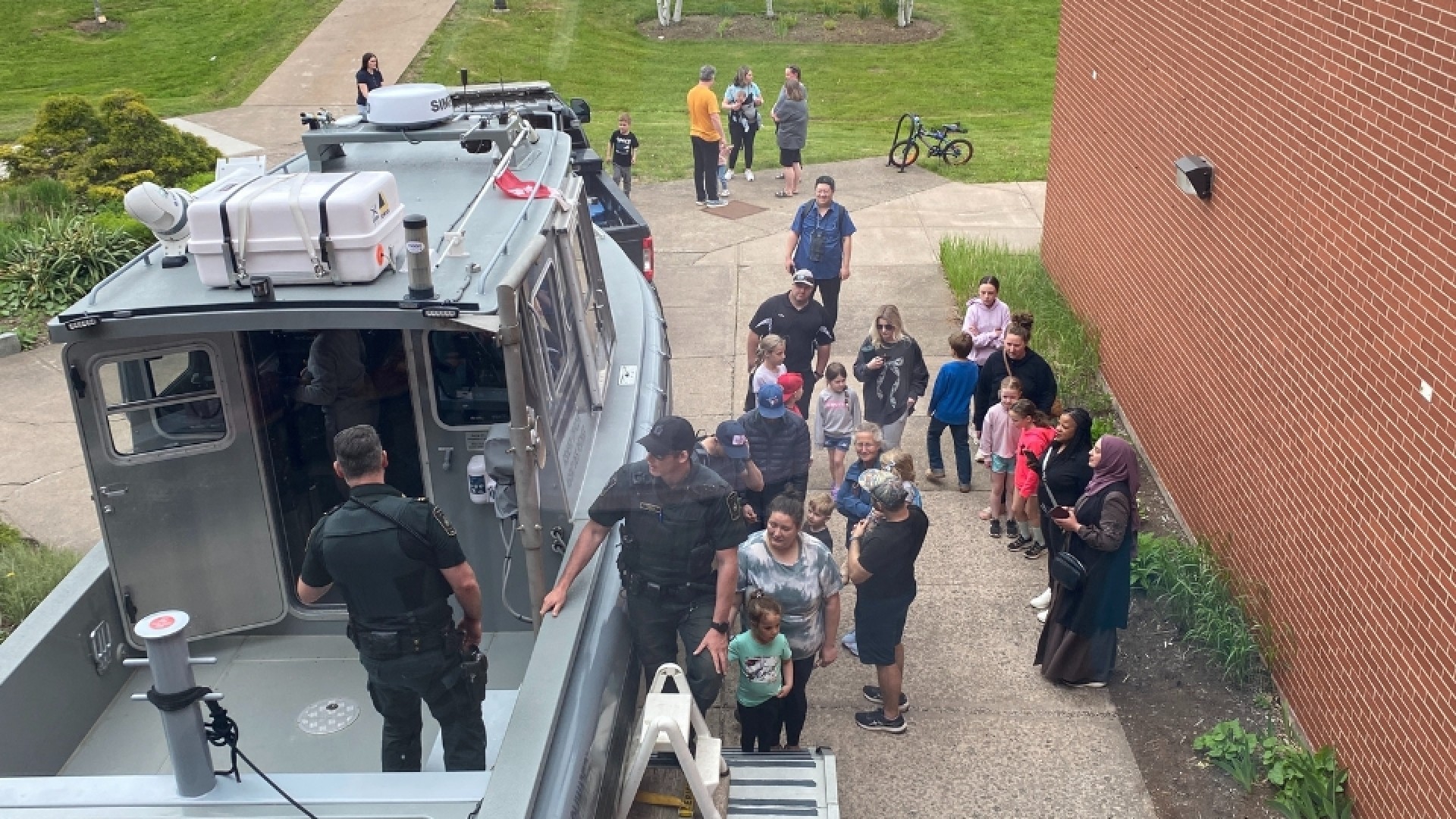 People gathered around a boat on a trailer near a brick building, with uniformed officers interacting with adults and children outdoors.