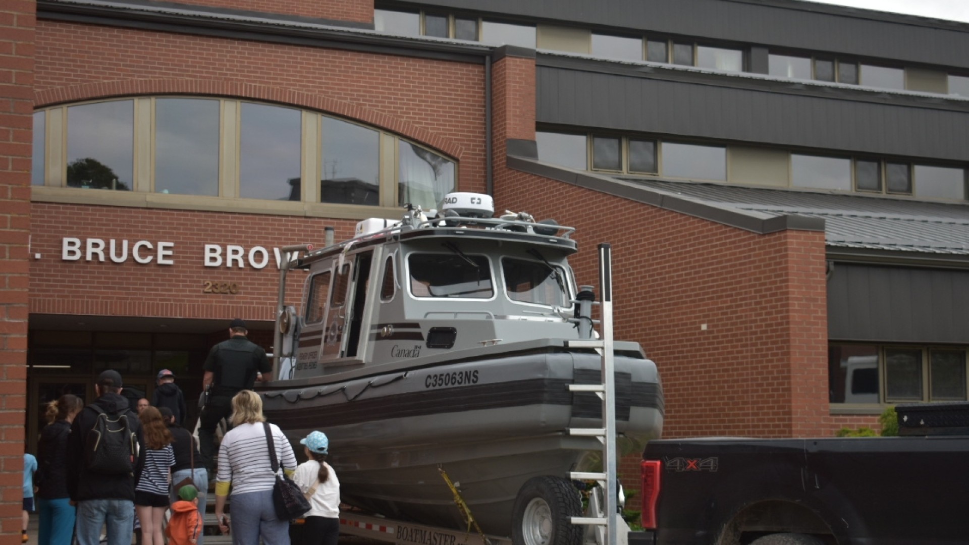 Children and adults lined up outside a building labeled 'BRUCE BROWN,' near a boat on a trailer hitched to a black pickup truck.
