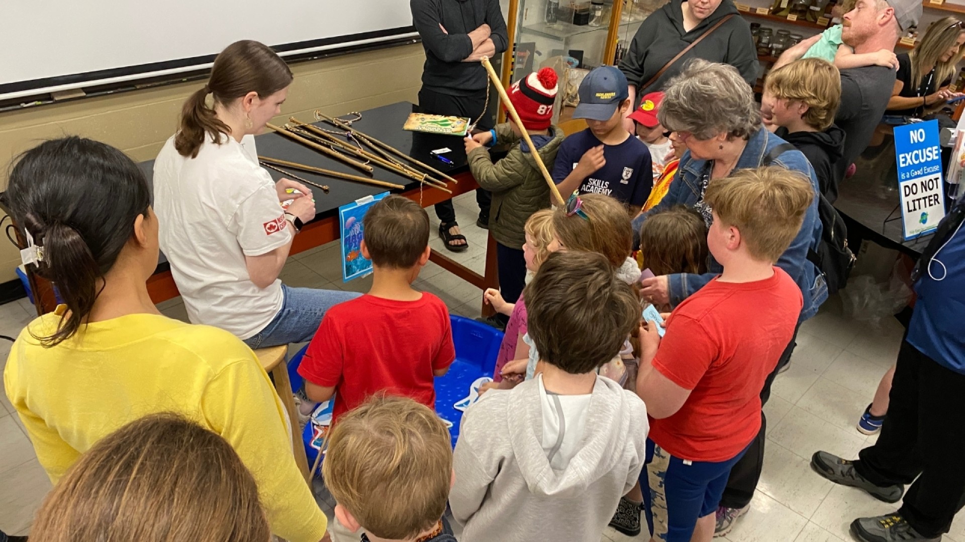 Children and adults gathered around a table with long sticks or rods in an educational setting, near a sign that reads 'NO EXCUSE IS GOOD ENOUGH TO LITTER.'