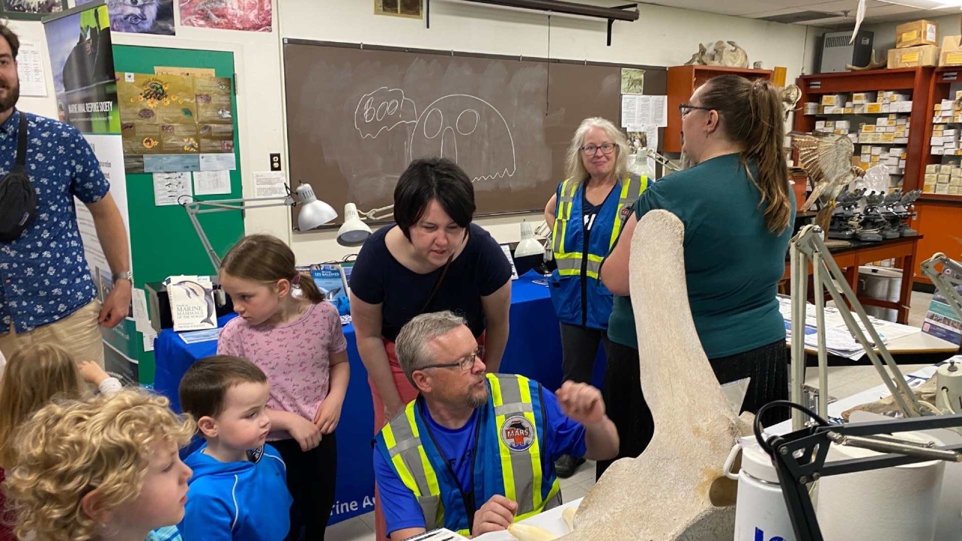 Children and adults gathered around a table with scientific displays, including a large bone or fossil, in an educational setting.