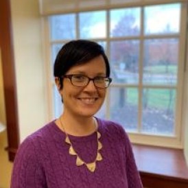Headshot of Sarah McKenna smiling in purple sweater in front of a window. 